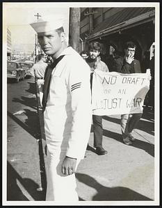 Bost Army Base Draft Protest Sailor waits for bus while protesters march behind him.
