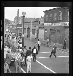 A crowd gathered in street near Dudley Door Window and Lumber and some running from a policemen approaching them