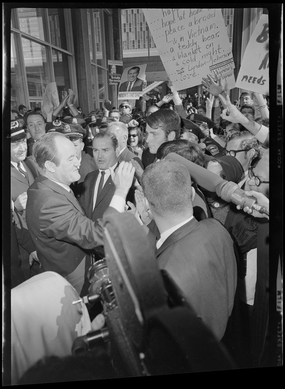 Hubert Humphrey walking through a crowd with people holding anti-Nixon and Lyndon Johnson posters