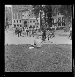 A shirtless man seated on the grass talking to another man with a bike