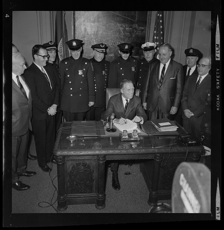 Mayor Kevin White at his desk signing Boston Police salary bill with officers behind him