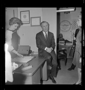 Mayor Kevin White sitting on a desk in City Hall as a woman flips through materials on his desk