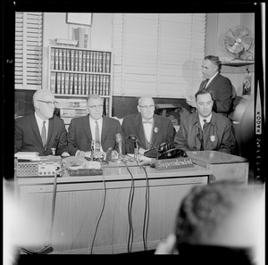 Four men seated at superintendent's desk, holding a news conference
