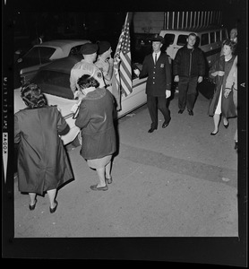 Officers and uniformed officials carrying a flag stand in front of cars