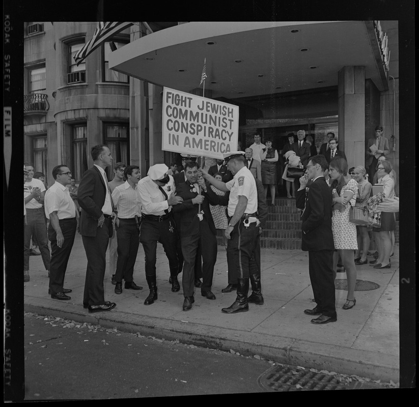 Police officers talking with a protester holding a sign reading "Fight Jewish Communist Conspiracy in America"