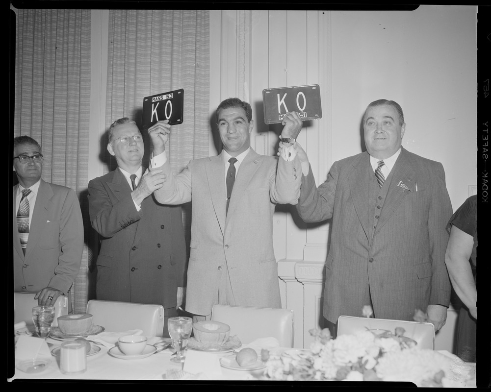 Rocky Marciano holds up new license plates that says "KO" while Governor Dever and Mayor Hynes look on
