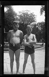 Two boys stand in front of a fountain wearing swimsuits