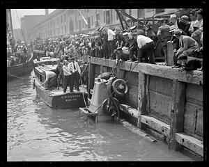 A crowd of people looking at an automobile being pulled out of the water next to a boat called the E.U. Curtis