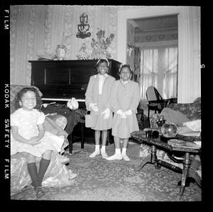Two girls stand by piano with another girl seated in chair