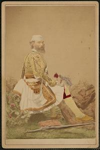 Bearded man in Grecian costume with rifle at feet