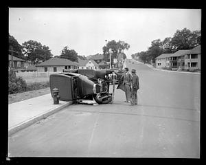 Two men stand examining an overturned car on its side