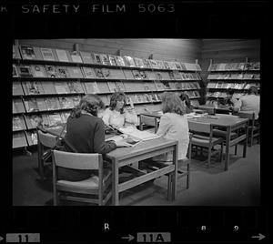 People sit at tables in a library, magazine display in background