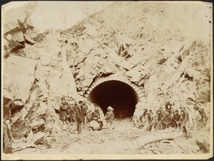 Group of men standing with tools (quarry hammers, etc.) in front of arched tunnel in mountain, possibly entrance to a mineshaft
