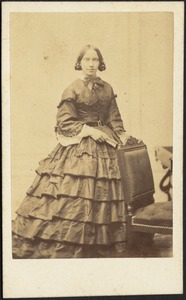 Woman in layered dress; standing near chair