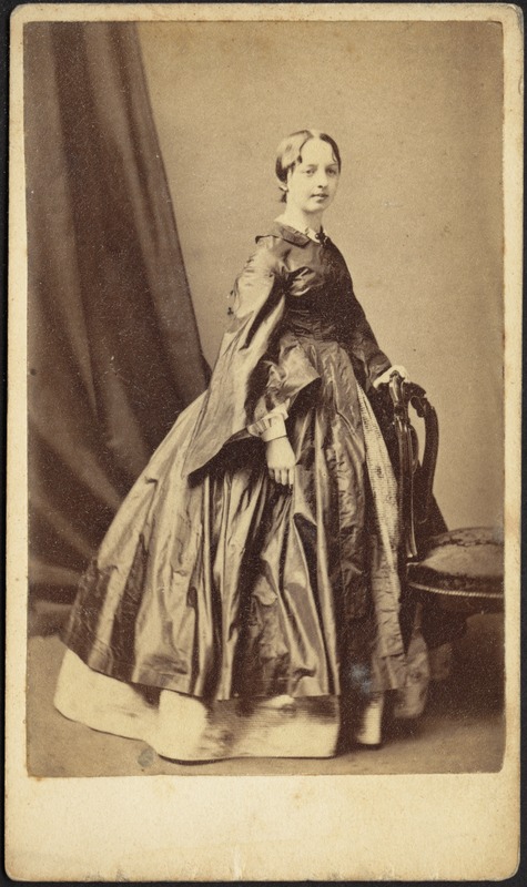 Young woman in satin dress with wide sleeves; standing near chair