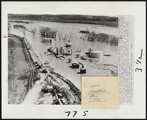 Oil Tanks Flooded; Workers Keep Battling -- While a group of oil tanks (center, right) stand surrounded by flooding water of the Missouri, levee workers (lower center) keep up the battle to continue building up dikes against the expected 31.5 foot crest. Trucks come down road (left) bringing supplies to the workers about three tiles north of here today.