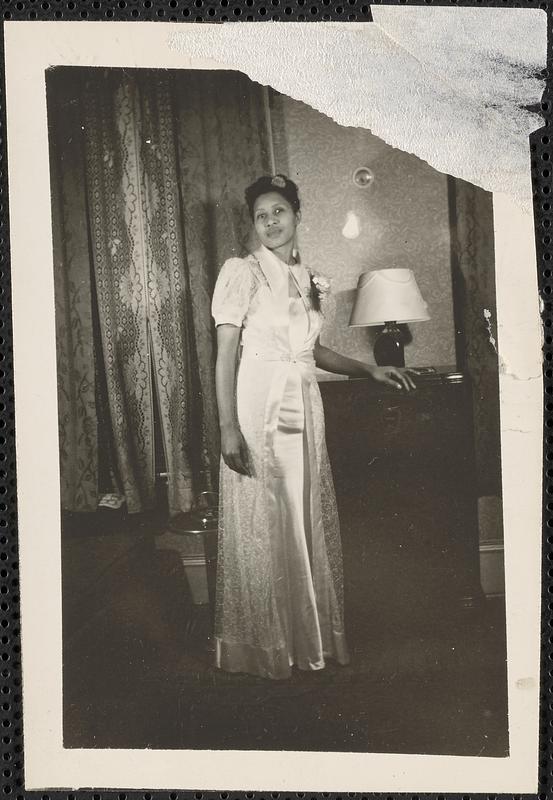 A woman in a floor length dress stands by a lamp