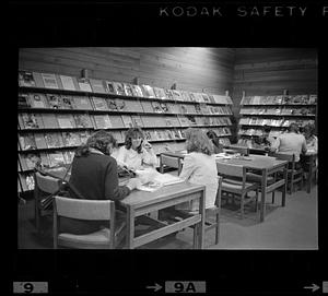 People sit at tables in a library, magazine display in background