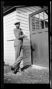 Young man wearing a hat and suit leans against a building