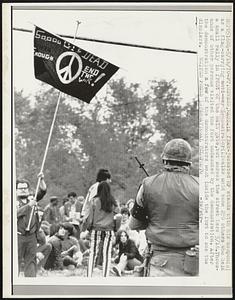 Ft. Devens, Mass.: A flak-jacketed MP stands with his weapon (R) as a flag-carrying anti-war demonstrator (L) walks amid some 250 others held a small rally in front of the main gate, but across the street here 5/16. Thousands of other persons visited the fort undaunted by the demonstration. After the demonstration a few of the demonstrators went inside the fort to see the displays.