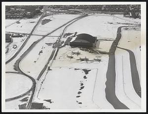 Snow-Bound Boston Airport and planes clustered on ground near American Airline's hangar. Plowed roads and taxiways etch patterns as seen in this aerial view. Upper left is East Boston expressway, football stadium and roads to airport. Right, wide taxi strips for planes.