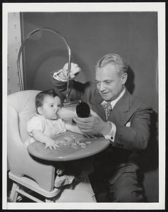 Bottle Holder. When N. W. Guden, above, became a grandfather two years ago, he invented a device which would aid in feeding the youngsters. This nursing bottle holder is patented and is being manufactured in Decatur, Ill. The device can be attached to a highchair or a crib and is adjustable. Guden says experiments have shown that the outer plastic chamber for the bottle serves to keep the liquids warm of cold. Here he is shown adjusting the gadgets as six-months-old Shayn Waddell partakes of the milk.