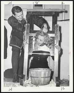 Carole Landis and Comic-Magician Russel Swann rehearse their guillotine act for the benefit of the Sixth War Loan Drive in New York. The 22-pound guillotine is one of Swann's famous props. To the innocent bystander the blade seems to be aiming at the head of the volunteer subject. Above, Swann adjusts Carole's head in the instrument. Below, Swann sends the blade of the guillotine down. Carole remains in one piece but a carrot that had been placed below her head is severed. Neat trick when it works.