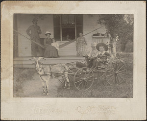 Adults and child on house porch, goat pulling children on carriage, c. 1900