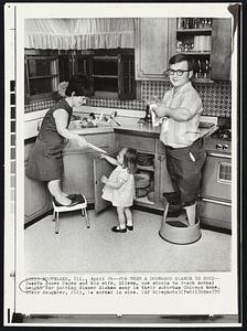 For them a Downward Glance is Good-- Dwarfs James Hagen and his wife, Eileen, use stools to reach normal height for putting dinner dishes away in their suburban Chicago home. Their daugther, Jill, is normal in size.