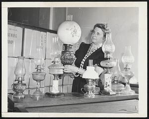 Lady Lamp Collector. Mrs. Emilie Brett is shown in her warehouse in downtown New York inspecting part of her collection of old-fashioned oil lamps ranging in age from 75 to 125 years. Mrs. Brett is the one-woman operator of the only firm in the country which supplies a "complete line of everything in kerosene lighting goods." The packing boxes in the background contain part of a 1000-gross shipment of cheap glass bowl oil lamp destined for Port of Spain.