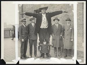 There’s some advantage in being tall, especially when one is in a crowd, says “Jim” Travers, the world’s tallest man. Jim stands 8 feet 9 inches and weighs 450 pounds. He is shown here with some normal-sized men, and also a midget, standing at his feet.