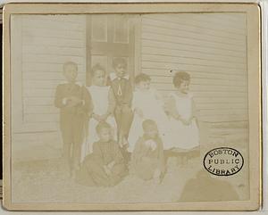Seven unidentified Black children, seated and standing outside the door of a clapboard building