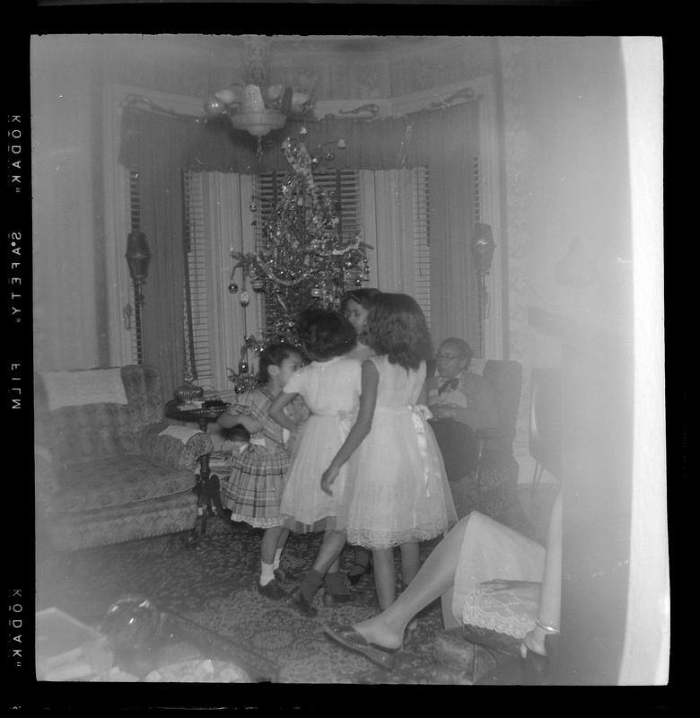 Girls in dresses stand in front of Christmas tree, a woman seated in an armchair in the background