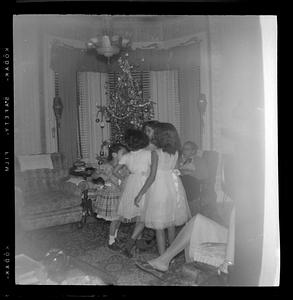 Girls in dresses stand in front of Christmas tree, a woman seated in an armchair in the background