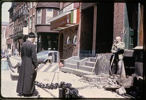 Two men, one wearing a cassock and a biretta, talk near Mamma Maria restaurant, North End