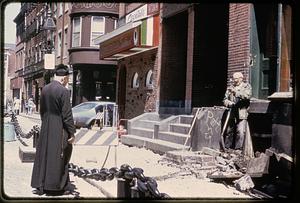 Two men, one wearing a cassock and a biretta, talk near Mamma Maria restaurant, North End