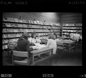 People sit at tables in a library, magazine display in background