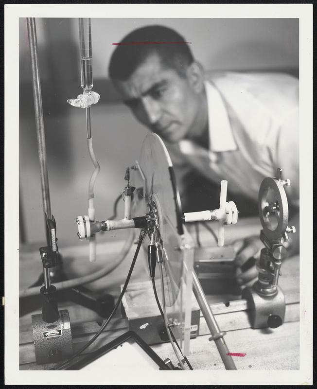 This liquid laser was developed in the Space Optics Laboratory at NASA'S Electronics Research Center in Cambridge. The part of it shown here is the ultrafast flash lamp. In the liquid laser a coaxial flash lamp surrounds the organic dyes which fill the cylindrical axis of the assembly. The energy storage capacitor is also mounted coaxially with the discharge system, and its electrodes are connected to the reflecting and conducting coatings applied on the outside surface of the outer quartz tube. This arrangement makes for the lowest possible conductance which results in very fast discharge. (Flash risetime is .05 micro-seconds, flash duration is .02 micro-seconds.) At the ends of the solution cell are placed reflectors which are broad band in the visible. In this manner the lasing output can be varied throughout the visible, from blue to red, continuously, merely by a simple and inexpensive changing of the dye solution itself. The scientist is Sejfi Protopapa.