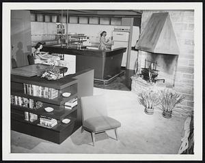 “New World Kitchen”. Designed by the General Electric Home Bureau, this 23 by 50-foot space is divided into three principal areas, a kitchen-laundry, dining room and living room. There are no walls between. It was constructed for showing at the International Home Furnishings Market held in Chicago. Pat Meehan, left, works a the electric ironer which is housed in the base of the counter when not in use, and Angel Casey is at the refrigerator-freezer behind the soda fountain counter.