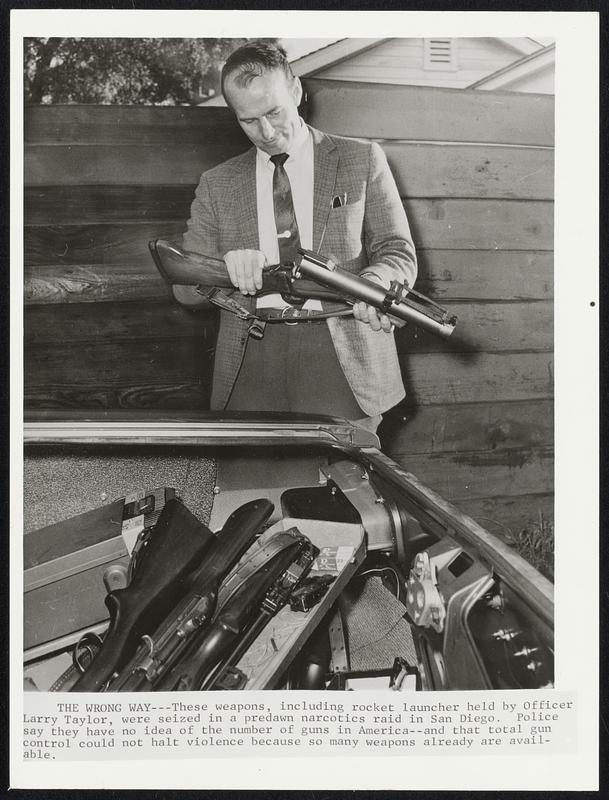 The Wrong Way--These weapons, including rocket launcher held by Officer Larry Taylor, were seized in a predawn narcotics raid in San Diego. Police say they have no idea of the number of guns in America--and that total gun control could not halt violence because so many weapons already are available.