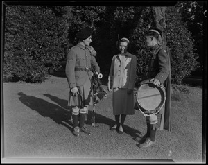 Two men wearing kilts and holding instruments speak to a woman