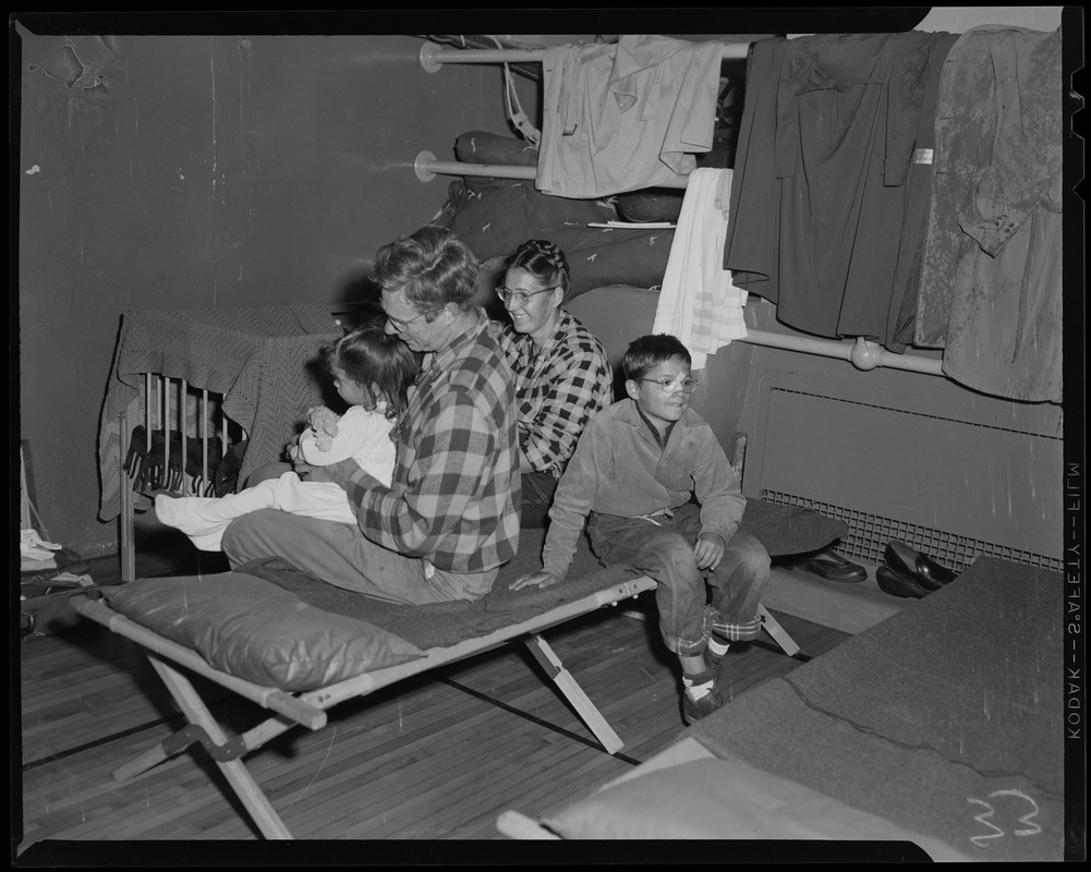 Family sitting on cots with clothing draped on pipes and a crib nearby ...