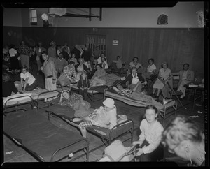 Children and adults on cots and folding chairs at a gymnasium shelter