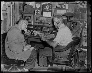 Men sitting at a desk, listening to a radio and telephone, taking notes