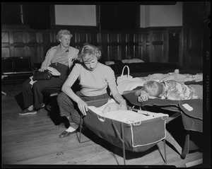 Woman sits on a cot at a shelter. She checks on her baby in a portable bassinet while another child sleeps beside her as another woman looks on