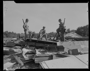 Two military men talking with a man and his dog, at the scene of destroyed homes and buildings