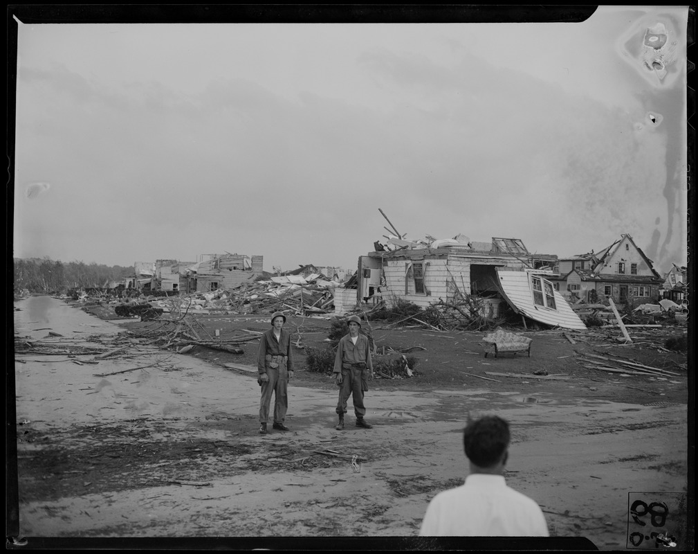 Two military men, standing in front of a string of destroyed homes ...