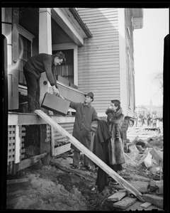 Man handing another man a suitcase from the porch of a damaged house