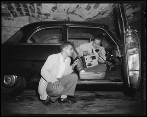 Two men, one in car and one kneeling in the street, using a radio instrument
