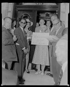 King Bhumibol Adulyadej and Queen Sirikit of Thailand standing by entrance to a building with a group of people holding a "Welcome Home to the King" sign